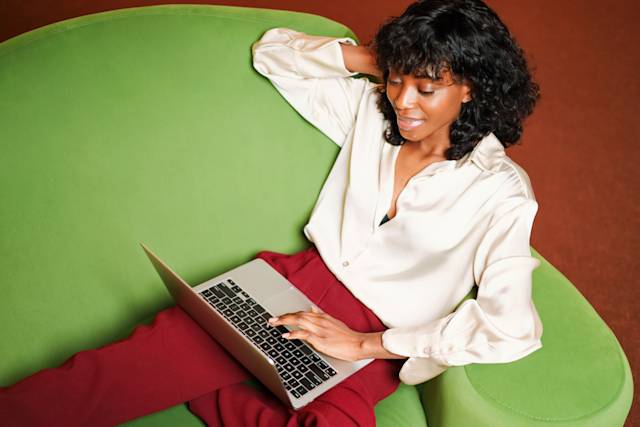 Woman in white blouse working on laptop while relaxing on bright green chair, wearing burgundy pants